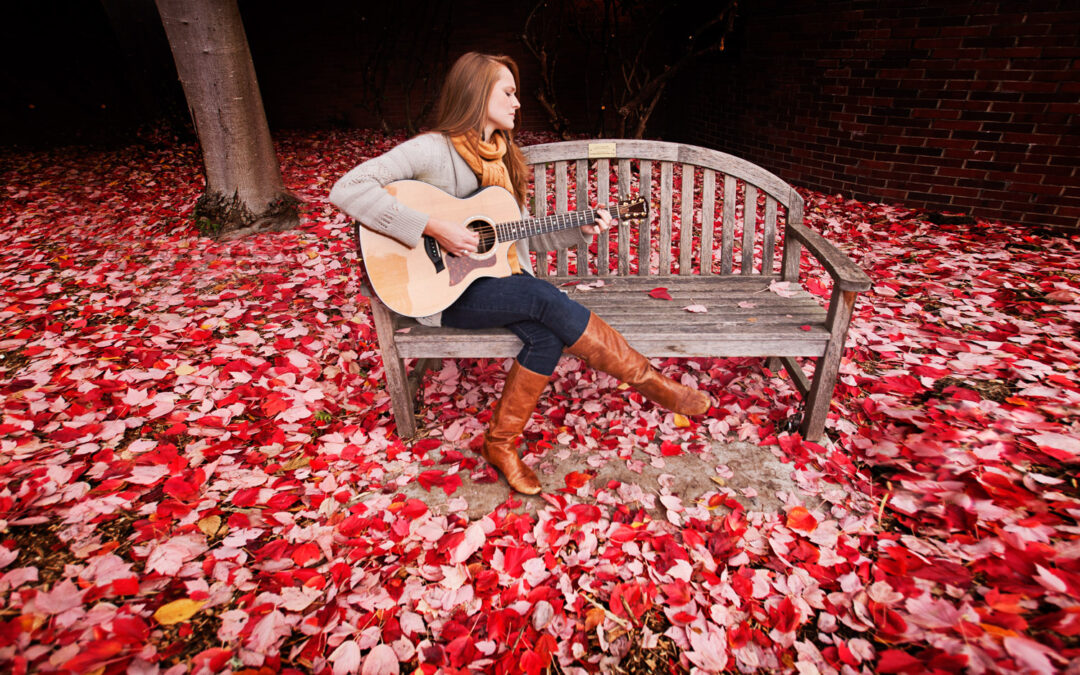 young woman playing guitar on bench surrounded by red leaves