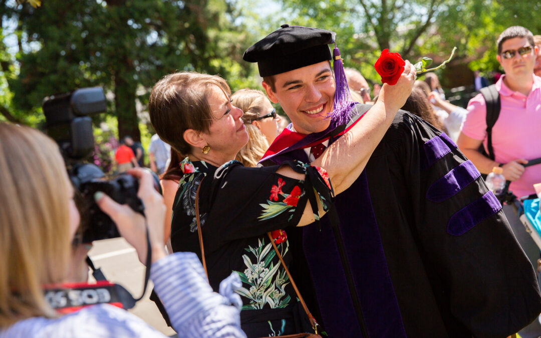 smiling student in graduation robe being hugged by woman with red flower